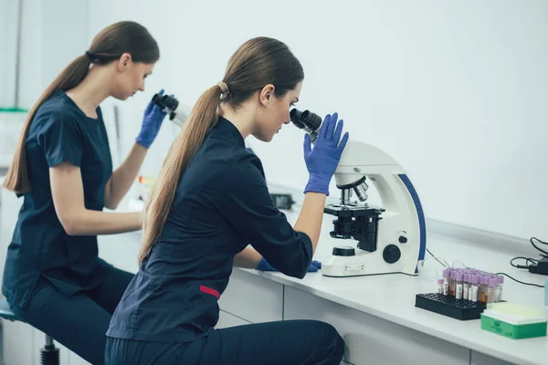 Two female lab scientists in dark blue uniform sitting at the table and looking through microscope lenses