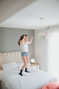 Güzel kız rahat kıyafetler ve kulaklık kulaklık kullanarak ve evde yatağında dans ederken gülümseyen müzik dinlemek