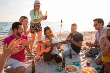 Happy multiracial young people playing guitar and making picnic at the beach. Friends are drinking beers and listening to music. Having fun at beach party in evening. clipart