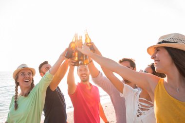 Şerefe! Bir grup mutlu genç sahilde bira şişeleriyle kadeh kaldırıyor. Kutlama ve tatil konsepti 