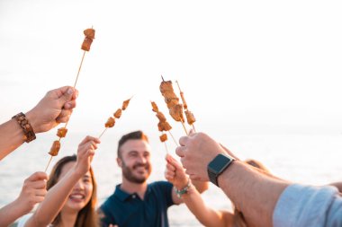 Happy friends having dinner a brochette that have been cooked in the barbecue on the beach. Summer time and holiday concept. clipart
