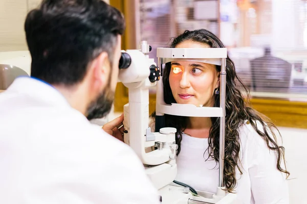 Ophthalmologist In Exam Room With Young Woman Sitting In Chair Looking Into Eye Test Machine