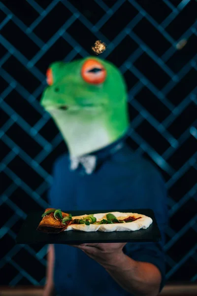Unidentifiable people waiter wears a Frog Head Mask showing mexican food on black background.