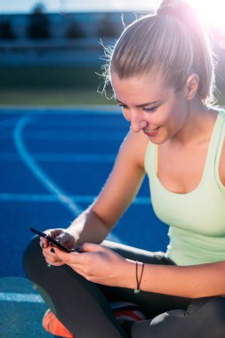 Mutlu genç atletik kadın mavi yarış pistinde çalıştırdıktan sonra siting süre Smartphone kullanarak.