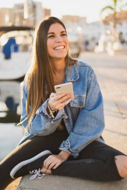 Bir smartphone kullanarak ve kameraya bakarak liman oturuyor ve gülümseyerek mutlu bir kadın