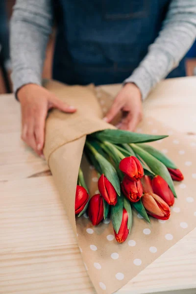Man florist makes red tulip bouquet and wrapping in pack paper on wooden table. Flowers