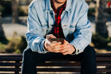 Parkta akıllı telefonunu kullanan güneş gözlüklü ve kot ceketli çekici bir genç.