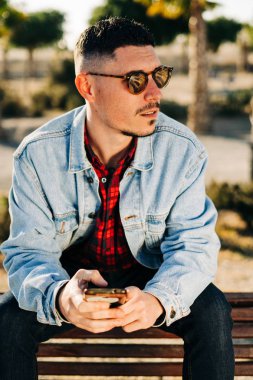 Parkta akıllı telefonunu kullanan güneş gözlüklü ve kot ceketli çekici bir genç.
