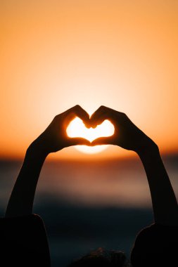 Dişi eller kalp şeklinde gökyüzüne karşı güneş ışınlarını uzatıyorlar. Sevgi dolu kalp şeklinde eller. Gün batımında Sevgililer Günü işareti.