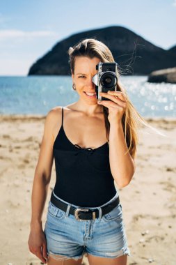 Yaz tatilinde plajda eski fotoğraf makinesiyle fotoğraf çeken çekici genç bir kadın.