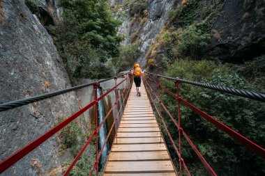 Turuncu sırt çantalı bir kadının asılı bir köprüden ormana gidişinin muhteşem görüntüsü..