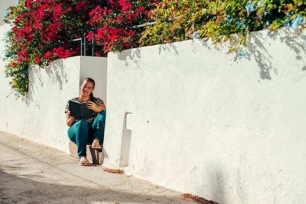 Gülümseyen genç kadın dışarıda kitap okuyor. Bir adım önde oturuyor. 