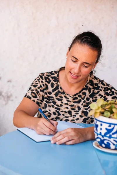 Woman write notes in her note book on blue desk outside, wearing animal print shirt.