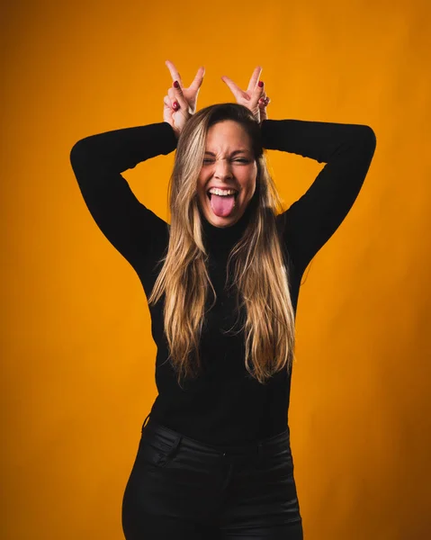 Menina loira bonita colocando as mãos sobre a cabeça enquanto se destaca a língua de uma forma engraçada e sorridente vestindo uma camisa preta e calças . — Fotografia de Stock