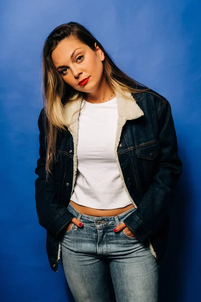Attractive young woman wearing white shirt, blue jacket, blue pants on which has his hands between the pocket with blue background. — Stok fotoğraf