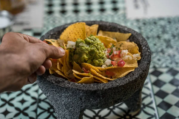 Hombre tomando chips de nacho de maíz amarillo adornado con carne molida, guacamole, queso derretido, pimientos y cilantro deja un molcajete, mortero tradicional mexicano . — Foto de Stock