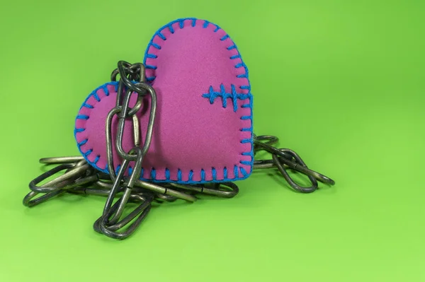 Heart-shaped trinket toy made of pink foam sheet and sewed with blue threads, connected to a metal chain and sitting on chroma key green surface background and viewed in close-up.