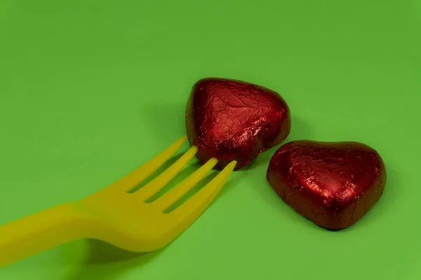 Two heart-shaped chocolate candies in red foil and yellow disposable plastic fork, viewed in close-up on green background. The concept of breaking hearts by womanizer or seducer and betrayal