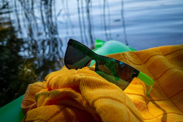 在黄色毛巾上的太阳镜放在漂浮在水面上的绿色充气床垫上 这是个暑假的概念 — 图库照片