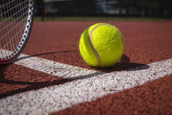 Tennis ball, line and racket on an outdoor court with the racket standing on end casting a shadow across the all weather surface