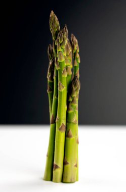 Bir grup taze, yeşil kuşkonmaz beyaz bir masada siyah bir arka plana karşı duruyor.