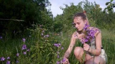 A beautiful light brown girl squatting on a blooming meadow collects a bouquet of wild flowers bells of a delicate purple color