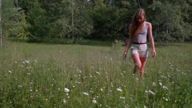 A beautiful light-brown girl walks through a blooming meadow, crouches down and collects daisies in an armful looks at the camera and smiles