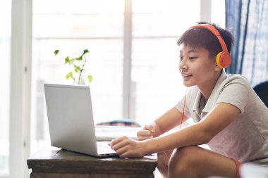 Asyalı çocuk evde online ders alıyor. Gülümsüyor ve kulaklık takıyor.