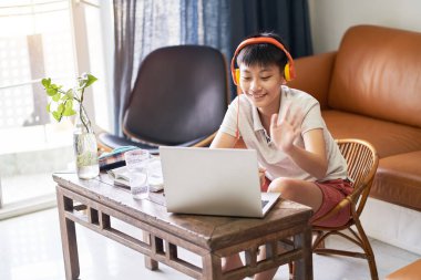 Asyalı çocuk evde online ders alıyor. Gülümsüyor ve kulaklık takıyor.