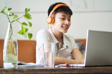 Asyalı çocuk evde online ders alıyor. Gülümsüyor ve kulaklık takıyor.