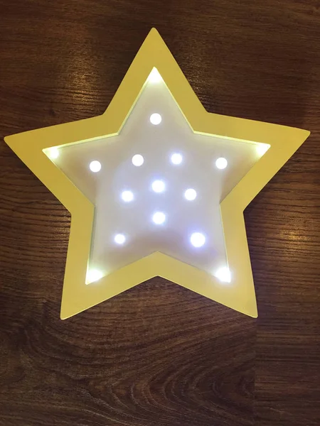 Glowing night light in the form of a star of a soft yellow color against a textured board made of dark brown natural wood