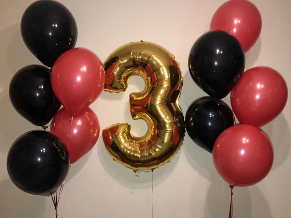 The composition of helium balloons in red and black, as well as a large golden number three. Decoration for a party dedicated to the celebration of three years