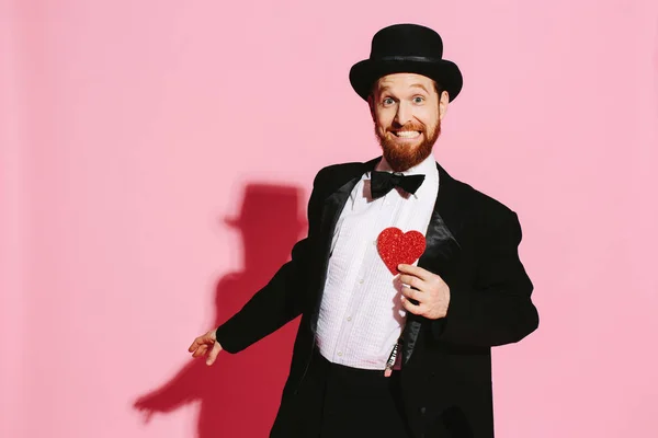 Smiling and dancing man in a tuxedo and top hat holding a red heart