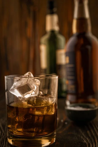 A glass of whiskey with ice, in the background are bottles on a wooden table of a bar counter, shallow depth of field, selective focus. The concept of alcoholic drinks in a roadside bar. — Stock Photo, Image