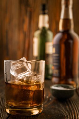 Arka planda buzlu bir bardak viski bir bar tezgahının ahşap masasında şişeler, sığ alan derinliği, seçici odaklanma. Yol kenarındaki bir barda alkollü içecekler konsepti.