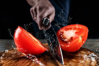 Şef büyük bir bıçakla kırmızı domatesi ikiye bölüyor, su ve meyve suyu farklı yönlere sıçrıyor ve havada donuyor, yüzeysel alan derinliği, seçici odaklanma, yemek pişirme konsepti.