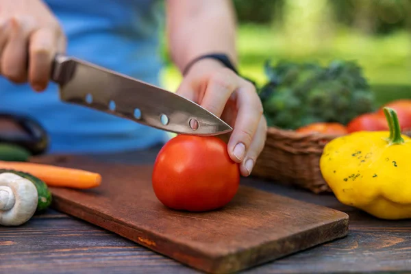 A woman is cutting vegetables, a womans hands are cutting a tomato on a salad on a wooden table, various other vegetables lie nearby. Healthy organic food concept.