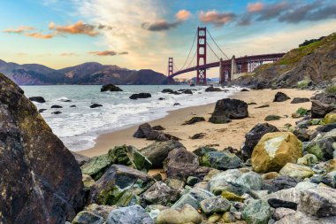 Golden Gate Köprüsü gün batımında Baker Sahili, San Francisco 'dan görüldü. Okyanus dalgaları, kayalar ve dramatik akşam gökyüzü manzarası. Seyahat, mimari ve macerayı simgeleyen ikonik Kaliforniya simgesi