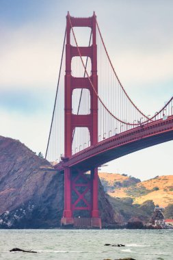 Golden Gate Köprüsü gün batımında Baker Sahili, San Francisco 'dan görüldü. Okyanus dalgaları, kayalar ve dramatik akşam gökyüzü manzarası. Seyahat, mimari ve macerayı simgeleyen ikonik Kaliforniya simgesi