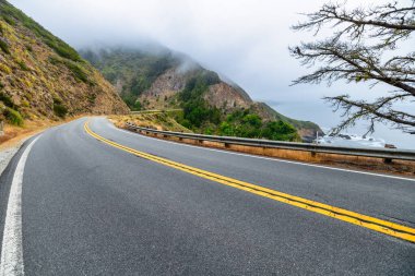 Winding Highway 1 road through the misty mountains of Big Sur, California. Scenic drive with rugged cliffs, pine trees, and coastal fog, capturing the dramatic atmosphere of this iconic route