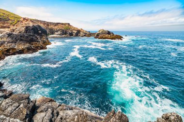 Dramatic Big Sur coastline along Highway 1, California. Waves crash against rugged cliffs, forming turquoise patterns in the Pacific Ocean, showcasing the raw beauty of the California coast