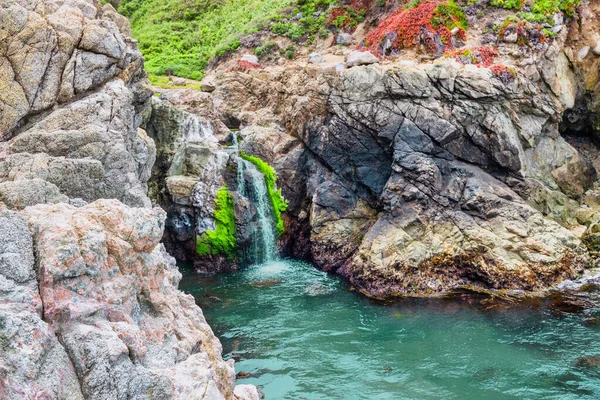 Small coastal waterfall cascading into a rocky tide pool along the Big Sur coastline, California. Green moss and colorful plants contrast with rugged cliffs, creating a hidden natural gem by the Pacific