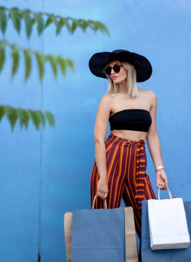 Şık genç kadın güneş gözlüğü, şapka ve pantolon bir şerit modaya uygun giyinmiş, bir açık hava Alışveriş Merkezi'nde alışveriş gider ve renkli bir arka plan üzerine poz veriyor