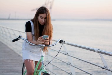 Güzel genç kadın bir bisiklet ile eğlenceli vardır, bir akıllı telefon kullanan, çağırır, sms, internet, sörf şafakta selfy yapar yazıyor