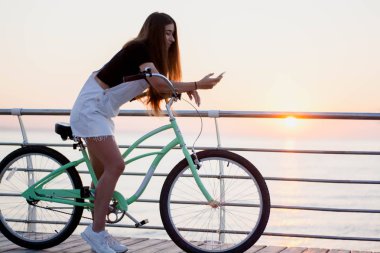 Güzel genç kadın bir bisiklet ile eğlenceli vardır, bir akıllı telefon kullanan, çağırır, sms, internet, sörf şafakta selfy yapar yazıyor
