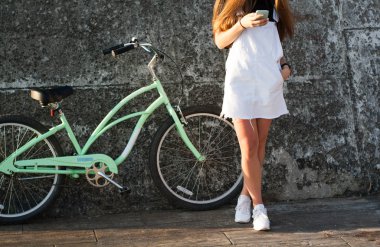 Güzel genç kadın bir bisiklet ile eğlenceli vardır, bir akıllı telefon kullanan, çağırır, sörf internet, sms, Güneş doğarken selfy yapar yazıyor