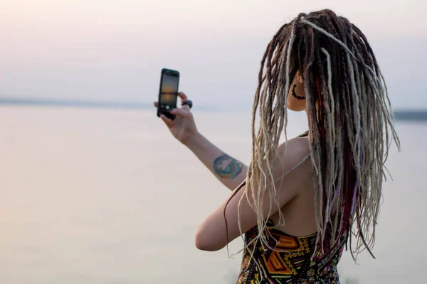 Genç güzel hippi kadın dreadlocks ile smartphone kullanır, doğa fotoğraflarını çekiyor ve bir selfie gün batımında alır
