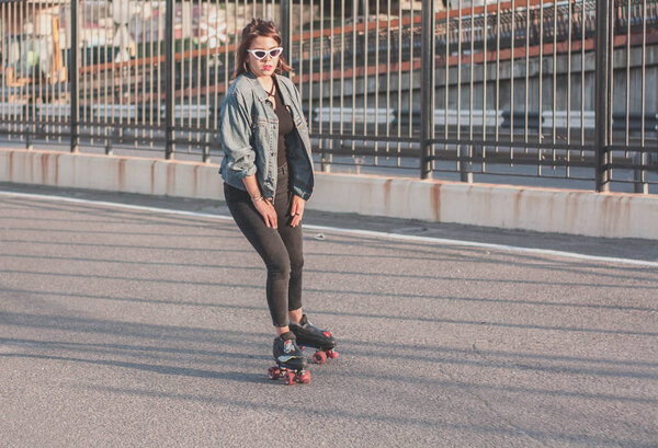 Stylish beautiful young woman in glasses and denim jacket rollerblading, dancing and having fun. Smiling and posing at sunset. Urban, rollers