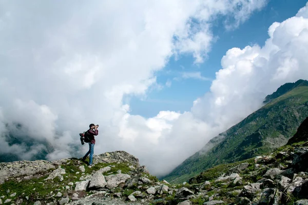 Tourist with a large backpack on his back stands on top of the mountain, holds an action camera in his hand and takes pictures