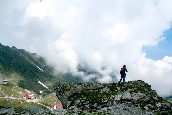 Tourist with a large backpack on his back stands on top of the mountain, holds an action camera in his hand and takes pictures